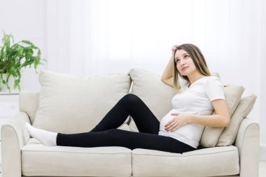 Young pregnant woman thinking about something on white sofa. Concept of pregnant woman.