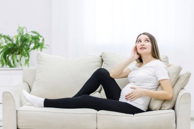 Young pregnant woman thinking about something on white sofa. Concept of pregnant woman.