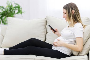 Young pregnant woman on white sofa using phone. Concept of pregnant woman.