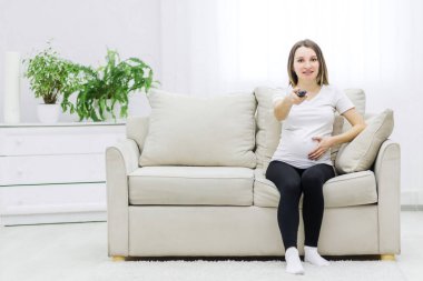 Cute pregnant woman sitting on sofa with remote control. Concept of pregnant woman with remote control.