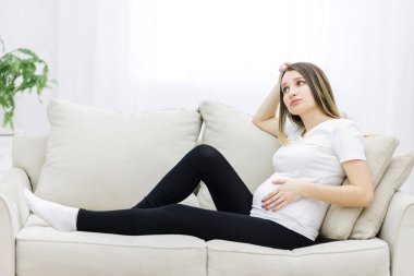 Young pregnant woman thinking about something on white sofa. Concept of pregnant woman.