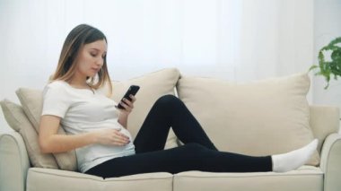 4k slowmotion video of pregnant woman sitting on white sofa with phone. Concept of pregnant woman.