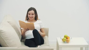4k video of pregnant woman on sofa with book and bowl of fruit on the table. Concept of pregnant woman.