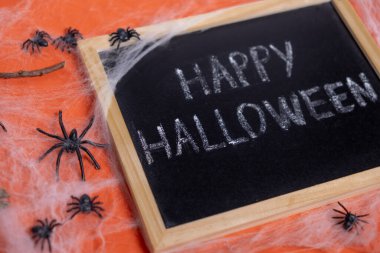 Turuncu arka planda Mutlu Cadılar Bayramı yazan bir karatahta. Örümcekler ağ örüyor 