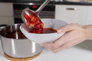 Ladling freshly cooked borscht with vegetables into a white bowl in a home kitchen setting clipart