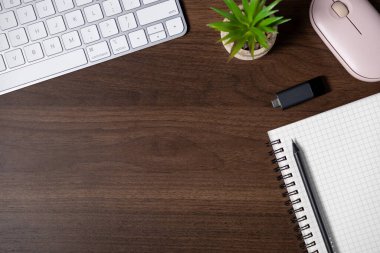 Çalışma alanının üst görünümü klavye, defter, kalem, USB sürücü, bitki ve pembe bilgisayar faresi ile ahşap masa üzerinde