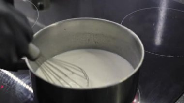 Ocağın üstünde paslanmaz çelik bir tencerede kremalı beyaz bir sos çırpmak, yakınlaştırma efektiyle yakın plan, pürüzsüz dokuyu vurgulamak.
