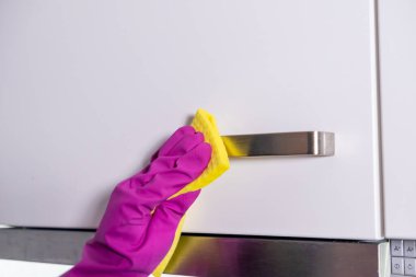 Beyaz mutfak dolabının sapını sarı bez ve pembe eldivenlerle temizliyorum. Hijyen ve parlaklık için.