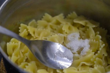 Adding salt to freshly cooked farfalle pasta in a metal pot with a spoon clipart