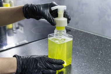 Mutfak tezgahında sarı sıvı sabun bulunan şeffaf sabun kutusu tutan siyah eldivenli eller, hijyen kavramı.