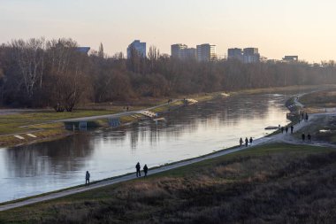 Huzurlu nehir manzarası huzurlu bir günbatımı akşamında yürüyüş yolları, ağaçlar ve uzak şehir silüeti. 