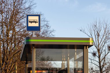 Bus stop sign on a modern shelter with green accents against a background of bare trees and clear sky during a sunny day clipart