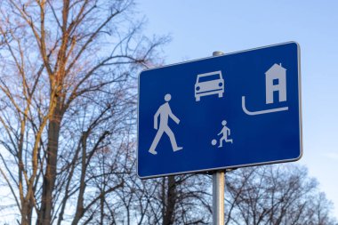 Üzerinde yaya sembolleri olan ikamet bölgesi trafik levhası, mavi arka planda top, araba ve evle oynayan çocuk, karışık kullanımlı bölge güvenliğini işaret ediyor.