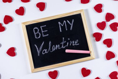 Beyaz tebeşirle yazılmış sevgililer günü karatahtam dağınık kırmızı keçeli kalplerle çevrili beyaz arka planda aşkı ve romantizmi simgeliyor.