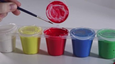 Canlı bir boya setinden fırçayı kırmızı boyaya batırmak, beyaz arka plandaki renkli konteynırlara yakından bakmak.