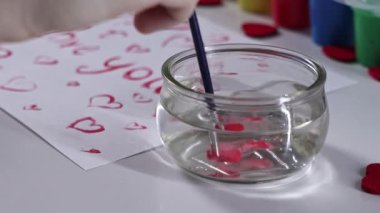 Cam bir kavanozdaki fırçayı suyla çalkalamak, kalp temalı dekorasyonlar ve sanatsal arka planla çevrili.