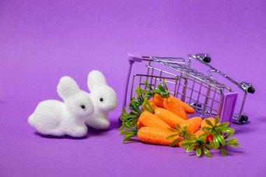 Paskalya dekorasyonu pelüş tavşanlar, parlak havuçlar ve mor arka planda ters dönmüş bir alışveriş arabası.