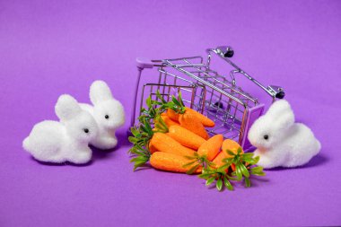 Paskalya temalı pelüş beyaz tavşancıklar, ışıltılı yapay havuçlar ve mor arka planda devrilmiş alışveriş sepeti.