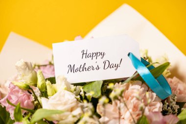 Zarif bir hediye etiketinin üzerinde anneler günü mesajı olan güzel bir çiçek buketi. Yumuşak mavi bir kurdeleyle süslenmiş. İçten ve sevgi dolu bir kutlama için sarı bir arka plana dizilmiş.