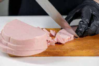 Olivier salatası hazırlığı için jambon dilimleme, ahşap bir tahtada taze sosis kesme, elleri siyah eldivenlerle eti tutma.