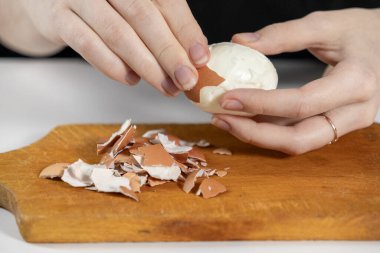 Yakın plan çekimde, ahşap bir kesme tahtasına haşlanmış yumurta kabuğu soyarken, etrafa saçılmış yumurta kabukları, ev yapımı yemek malzemeleri hazırlarken.