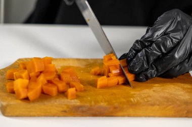 Siyah eldivenli şef, profesyonel mutfak ortamında keskin bir bıçakla salata hazırlamak için tahta kesme tahtasının üzerinde haşlanmış havucu küçük küplere ayırıyor.