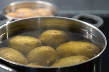 Fırının üstünde paslanmaz çelik bir tencerede patates haşlarken yemek hazırlamak için arka planda kaynayan başka bir yumurta tenceresiyle birlikte pişerken buhar çıkıyor.