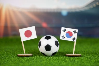 Japonya, Güney Kore futbol maçında bayraklar, yeşil sahada futbol topu ve bulanık stadyum, Asya spor rekabetini ve takım çalışmasını sembolize ediyor.