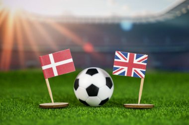 Danimarka ve Birleşik Krallık futbol müsabakaları ulusal bayraklar, yeşil sahada futbol topu ve stadyum arka planı ile rekabetçi spor ruhunu temsil ediyor.
