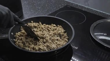 Elektrikli sobada kızartma tavasında et pişirmek, eldiven giyerken siyah spatulayla karıştırmak, sıcak bir yüzey üzerinde ev yapımı kıymalı et pişirmek.