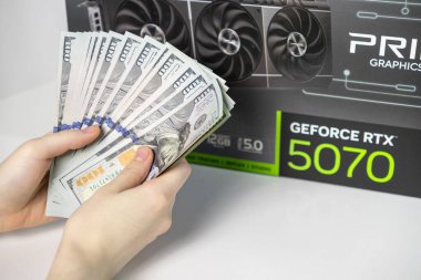 Poznan, Polonya - 28 Mart 2025: Asus GeForce RTX 5070 kutusunun önünde üst düzey modern grafik kartlarının maliyetini gösteren ABD doları destesi tutan kişi