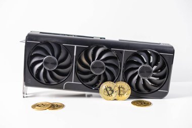 Fiziksel Bitcoin paralarıyla çevrili beyaz bir yüzeyde siyah üçlü fan GPU. Modern kripto para birimi madenciliğini ve dijital finansı temsil eden kavramsal görüntü