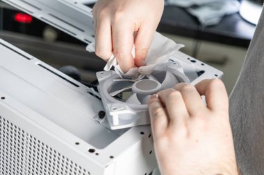 Bir teknisyen bilgisayar bakımı sırasında beyaz bir bilgisayar soğutma vantilatörünü bez kullanarak temizliyor. Vantilatör açık bir davanın üst paneline yerleştiriliyor ve toz temizleme işlemini gösteriyor.