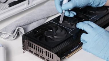 Mavi eldiven giyen teknisyen pamuklu bez kullanarak bir grafik kartının soğutma vantilatörünü dikkatlice temizliyor. Bakım ve toz önleme sahnesi