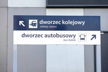 Modern gri duvar arka planında polonyaca ve İngilizce dilinde ok ve simgelerle tren istasyonu ve otobüs durağına giden yolu gösteren yön işareti