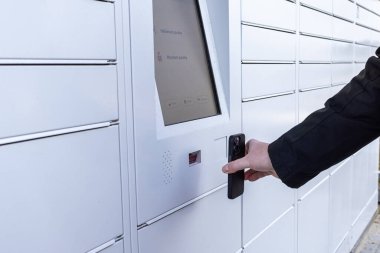 Beyaz parsel dolap ekranındaki QR kodunu taramak için akıllı telefonu kullanan bir adam şehirdeki sokaktaki bağlantısız dağıtım sistemiyle paketi almak ya da göndermek için