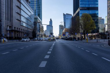 Varşova, Polonya - 3 Nisan 2025: Uzun cam binaları, trafikte arabaları ve kaldırımlarda yürüyen insanları olan modern şehir caddesi. Açık mavi gökyüzünün altında şehir silüeti