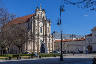Varşova, Polonya - 3 Nisan 2025: Polonya 'nın Varşova şehrinin tarihi bölümünde süslü cepheleri ve heykelleri olan güzel barok kilisesi. Parlak güneşli hava ve meydanda yürüyen insanlar.