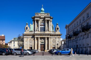Varşova, Polonya - 3 Nisan 2025: Orta Varşova, Polonya 'da sütunları, heykelleri ve yeşil kubbeleri olan barok tarzı kilise cephesi. Güneşli bir günde insanlar yürüyor ve arabaları ön tarafa park ediyor.
