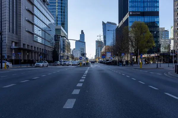 Varşova, Polonya - 3 Nisan 2025: Uzun cam binaları, trafikte arabaları ve kaldırımlarda yürüyen insanları olan modern şehir caddesi. Açık mavi gökyüzünün altında şehir silüeti