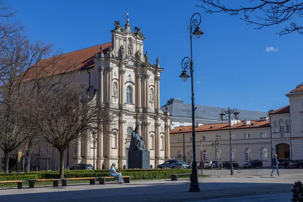Varşova, Polonya - 3 Nisan 2025: Polonya 'nın Varşova şehrinin tarihi bölümünde süslü cepheleri ve heykelleri olan güzel barok kilisesi. Parlak güneşli hava ve meydanda yürüyen insanlar.
