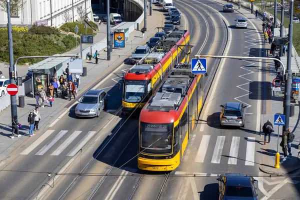 Varşova, Polonya - Nisan 03, 2025: Polonya 'nın Varşova şehrinde, yayalar, arabalar ve güneşli bir günde bir tramvay durağı ile çevrili yoğun bir caddede iki modern tramvay seyahat ediyor.