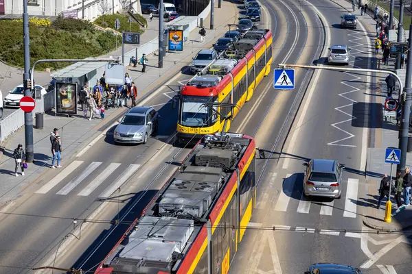 Varşova, Polonya - Nisan 03, 2025: Polonya 'nın Varşova şehrinde iki kırmızı ve sarı tramvay, yakın araçları, tramvayı ve yayaları olan hareketli bir cadde boyunca parlak bahar güneşi altında seyahat ediyor.