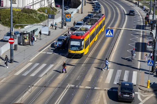 Varşova, Polonya - 3 Nisan 2025: Polonya 'nın orta kesimindeki bir yaya geçidinde kırmızı ve sarı bir tramvay duruyor. İnsanlar yürür, bisiklet sürer ve güneşli bir günde kalabalık bir cadde boyunca bekler.
