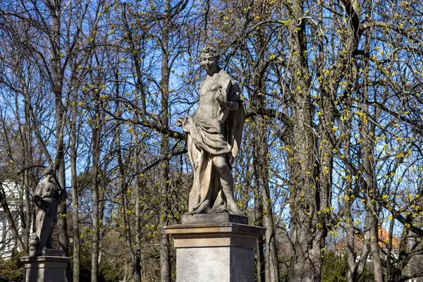 Varşova, Polonya - Nisan 03, 2025: Akan giysilerle süslenmiş klasik bir erkek figürünün taş heykeli, huzurlu bir park ortamında tomurcuklanan yapraklarla ilkbahar ağaçları arasında bir kaidede duruyor.