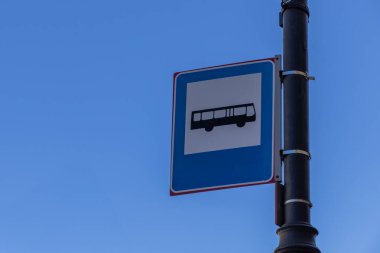 Bulutsuz mavi gökyüzüne karşı siyah bir direğe kurulmuş otobüs durağını gösteren minimalist toplu taşıma levhası. Temiz ve sembolik kentsel navigasyon ögesi