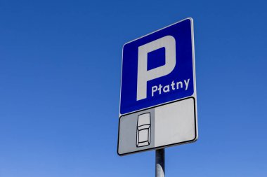 Araçlar için ücretli park yerini gösteren yol tabelasının yakın çekimi. Mavi ve beyaz tabela bir araba sembolü içeriyor ve açık mavi gökyüzüne ayarlı.
