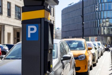 Şehrin caddesinde arka arkaya birden fazla araba park etmiş bir parkmetreye yakın çekim. Binaları ve güneş ışığıyla modern kentsel çevre