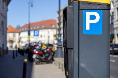 Şehir parkmetresine mavi bir parkmetre yakın plan. Arka planda, motosikletler ve arabalar güneşli bir şehir caddesi boyunca park halindeler.