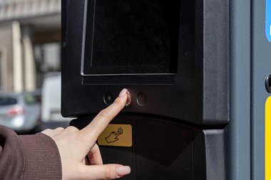 Bir kadının eli dijital parkmetrede dokunmatik ekranlı bir düğmeye basar. Cihaz bir şehir caddesinde gün ışığında bulunuyor.